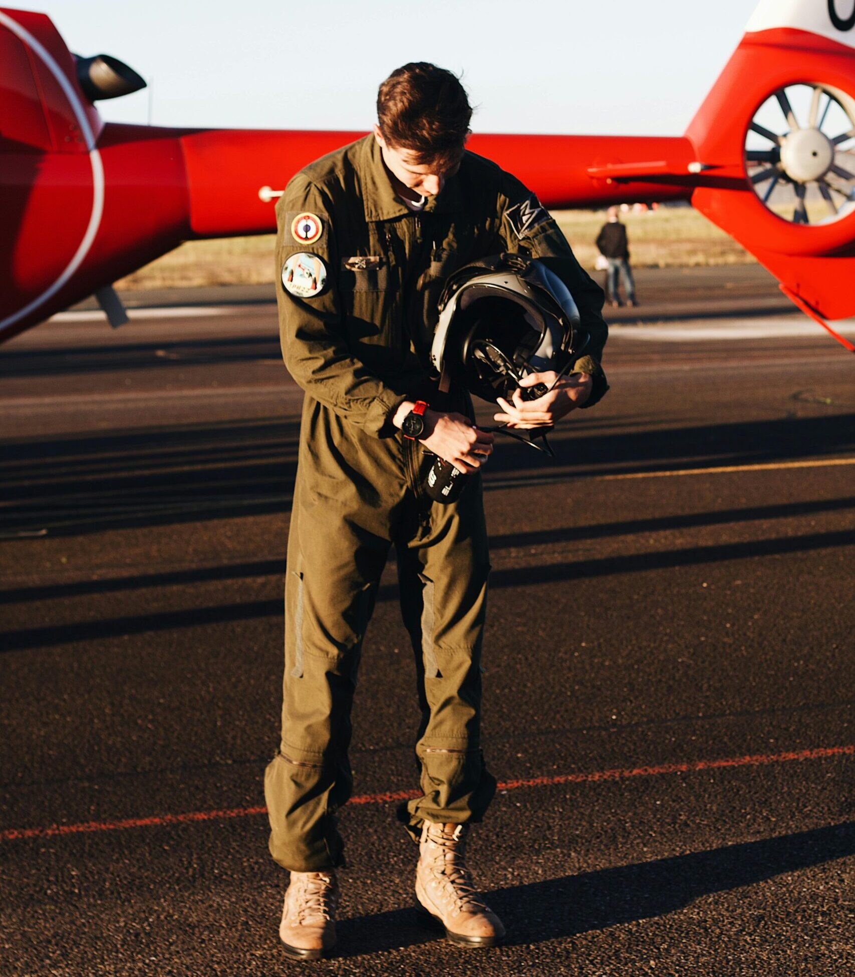 A pilot holding a helmet, standing by a red aircraft on a clear day at the airport.